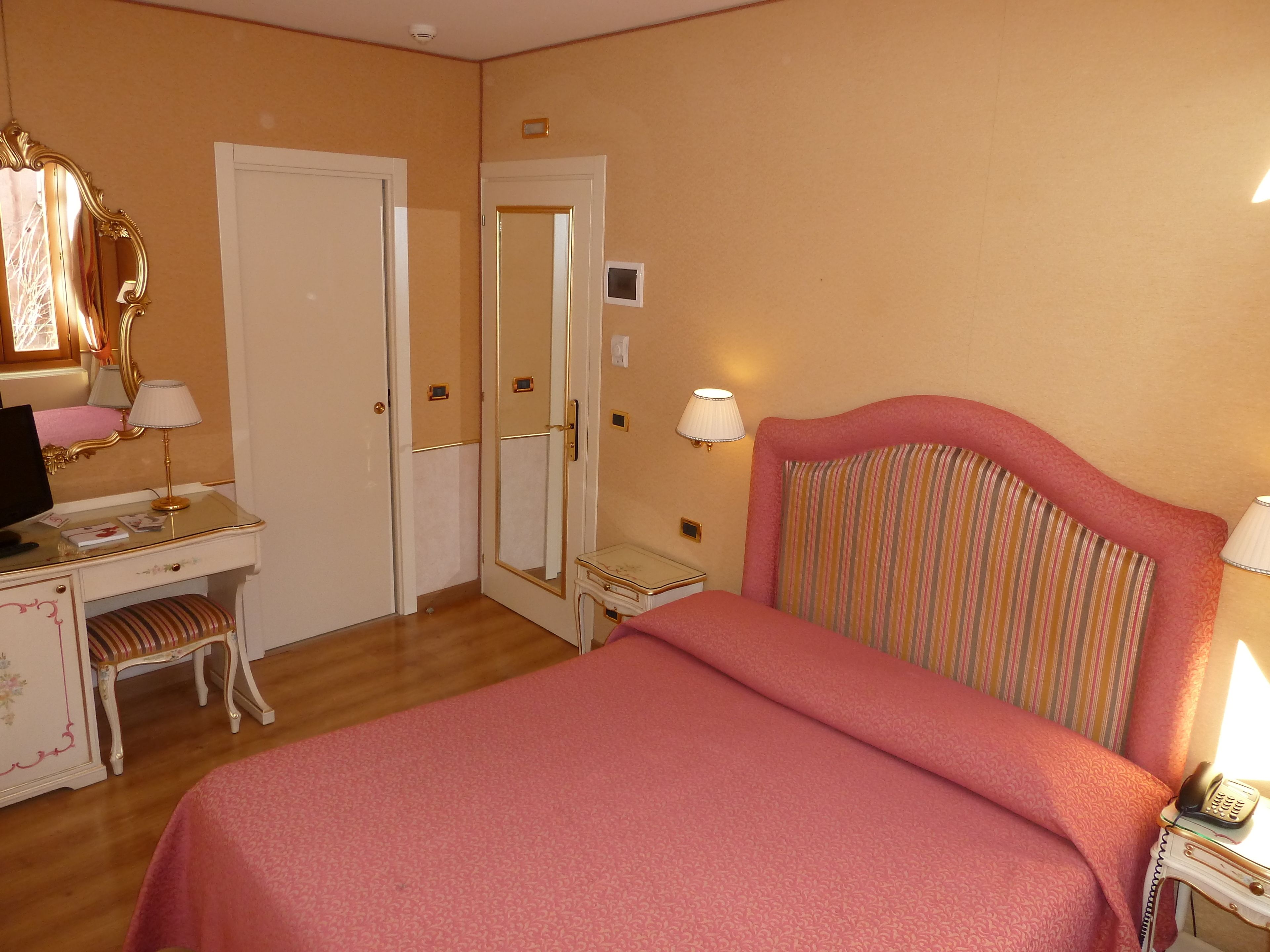 Una camera d'albergo con un letto, una scrivania con una sedia, uno specchio e due lampade da comodino.