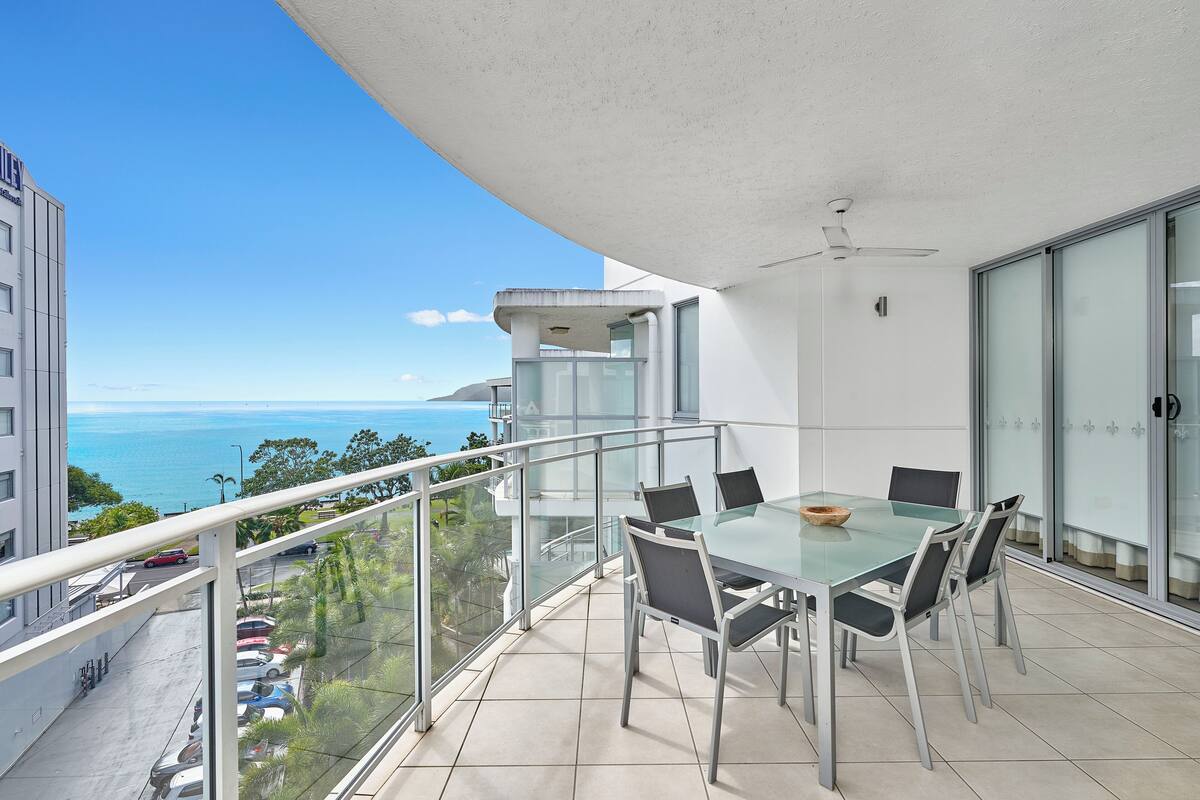 A balcony with a glass table and chairs, overlooking a parking area and a view of the ocean.