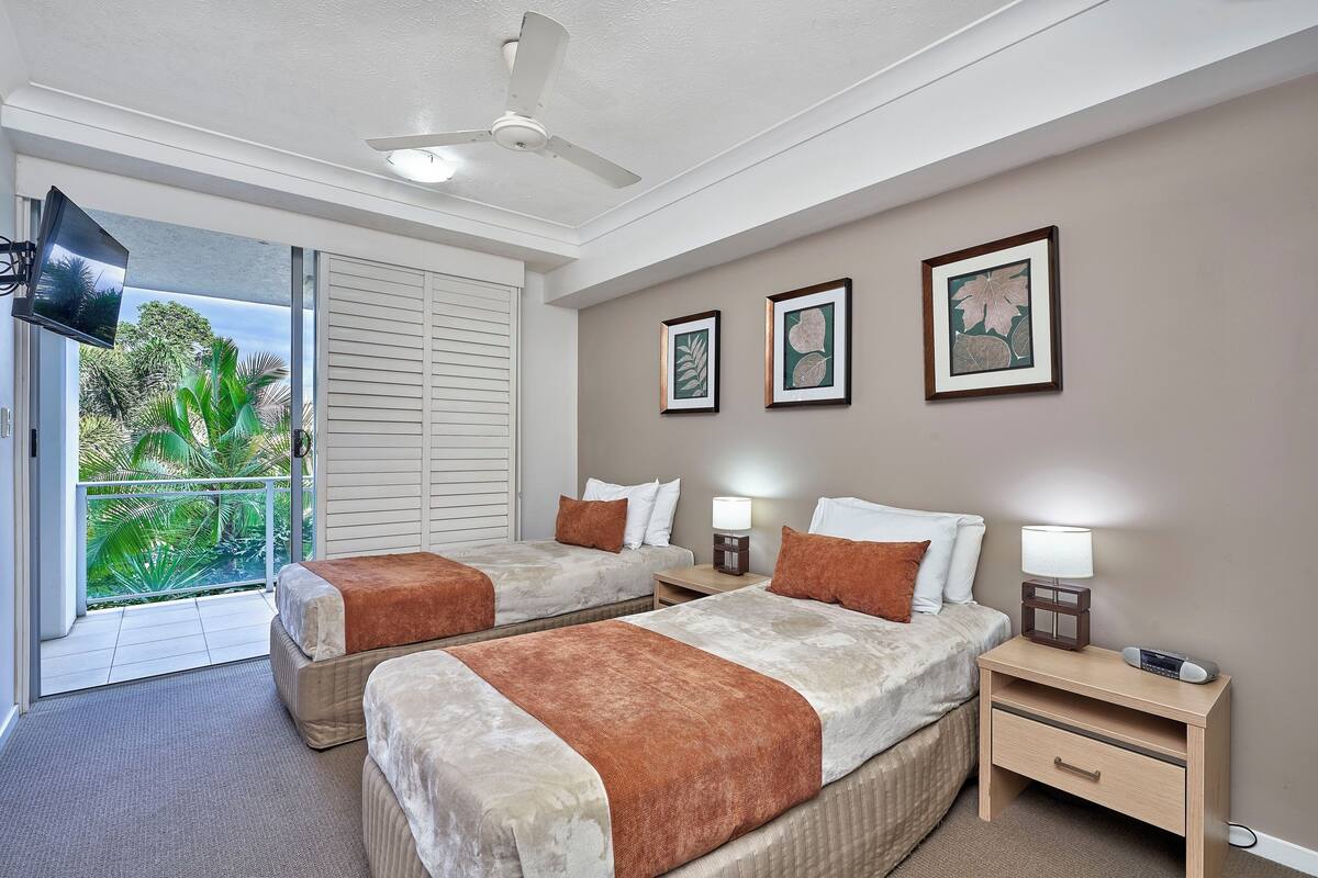 A hotel room with two beds, a ceiling fan, a TV, framed artwork, a nightstand with a lamp, and a view of greenery through the balcony doors.