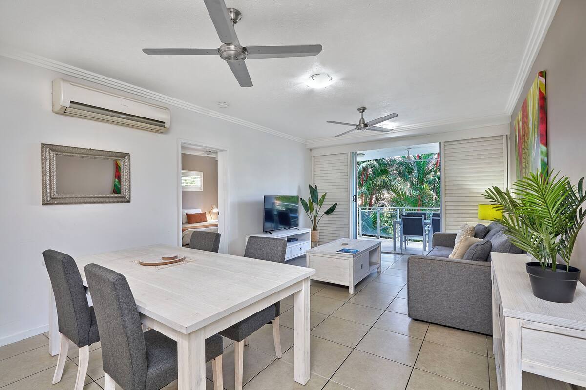 A modern living room with a dining area, two ceiling fans, a TV, and a balcony with plants.