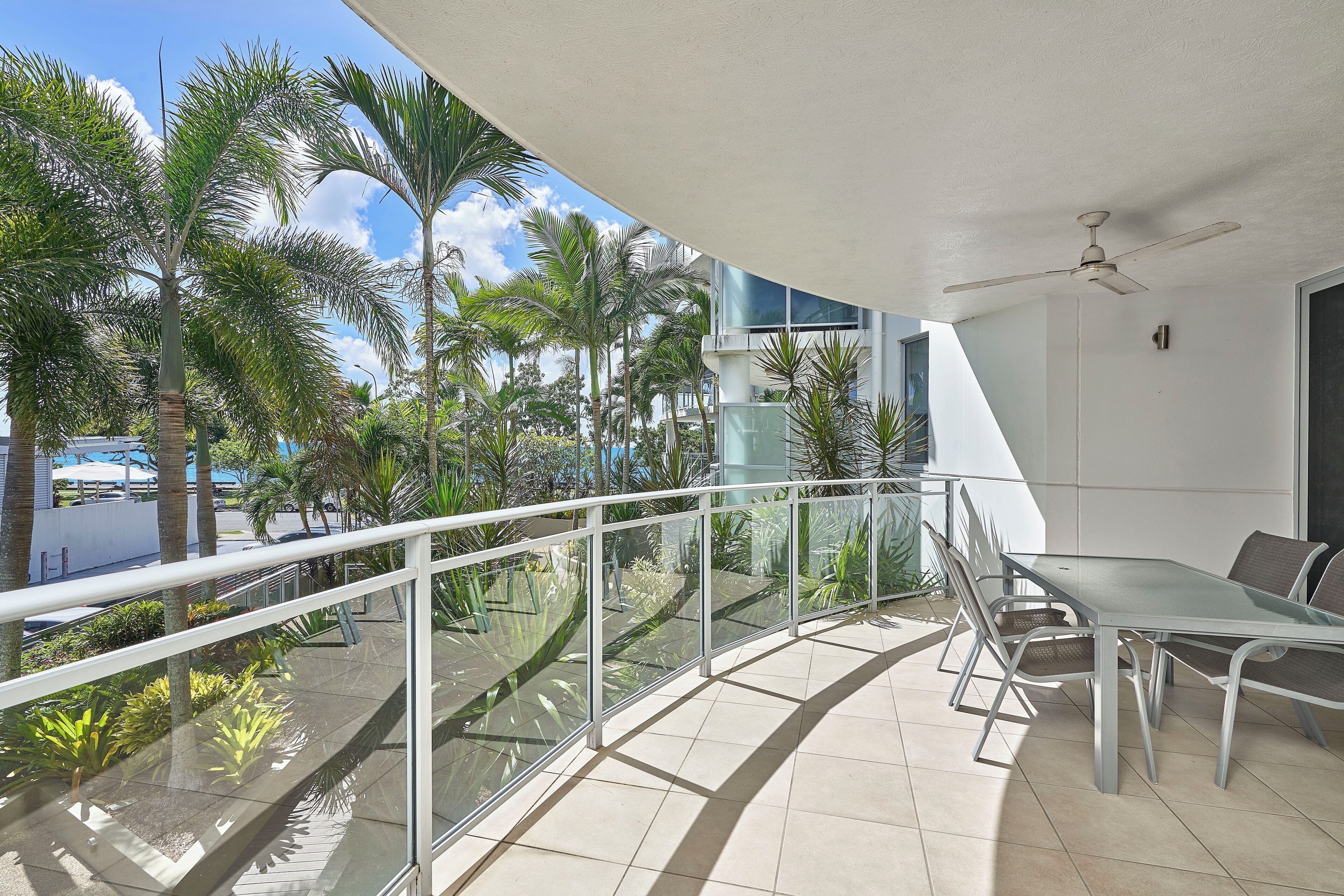 A balcony with a glass railing, a table, and chairs, overlooking a palm tree-lined area.