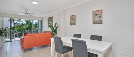 Uma área de jantar moderna com uma mesa branca e cadeiras cinzas, um ventilador de teto e vista para uma varanda com plantas.