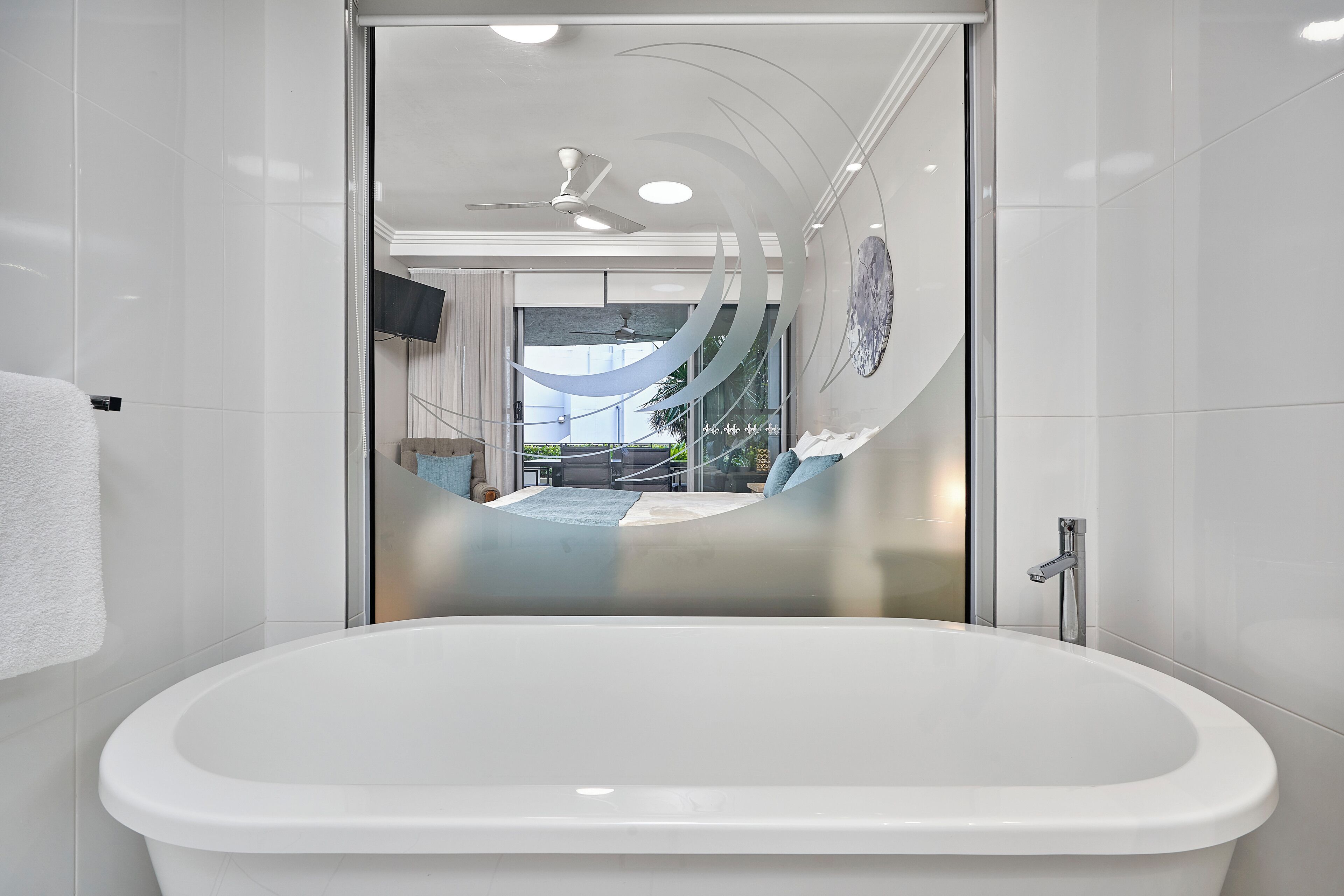 A modern bathroom with a freestanding bathtub, a glass shower enclosure, and a visible living area with a sofa and a TV.