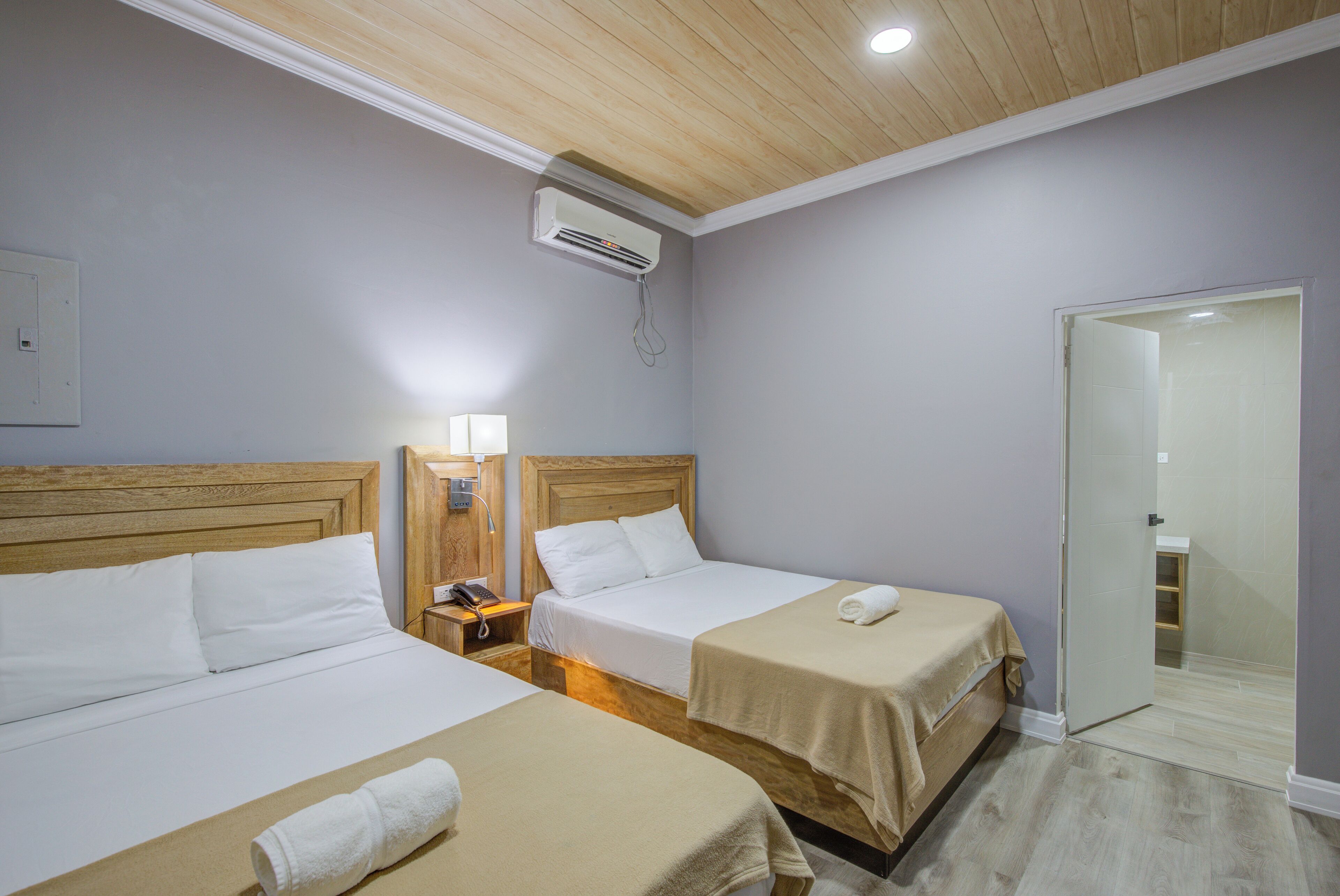 Una habitación de hotel con dos camas, techo de madera, unidad de aire acondicionado, lámpara de noche y puerta de baño.