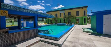 A swimming pool area with a blue pool, a covered seating area, and a building with a green roof.