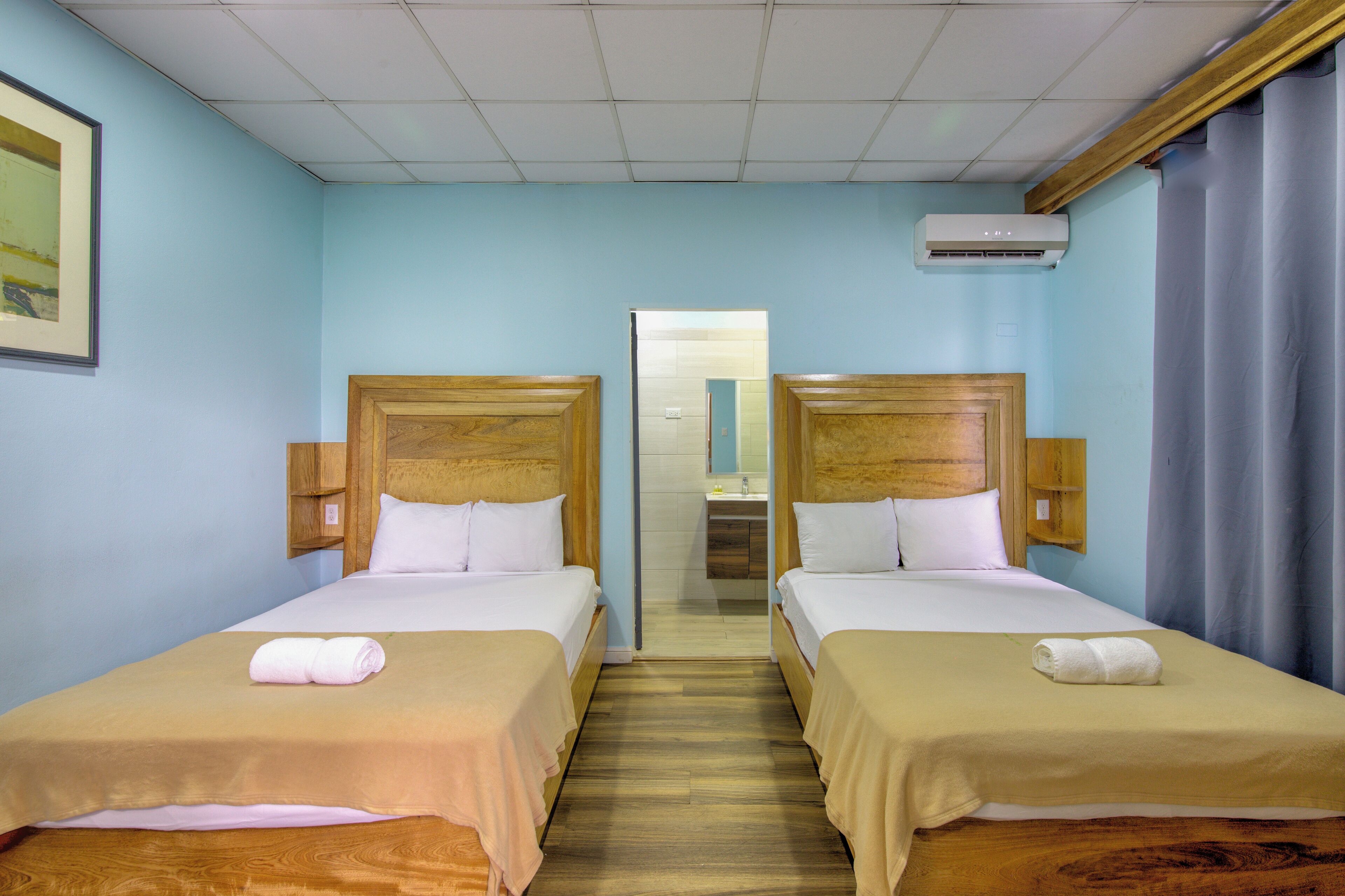 A hotel room with two single beds, wooden headboards, beige bedding, a wooden floor, a framed picture on the wall, and an air conditioning unit.