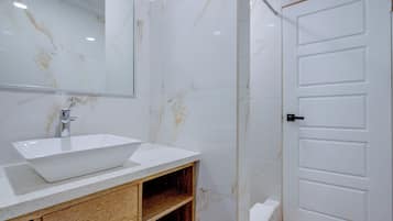 Un baño con lavamanos blanco, tocador de madera, paredes de mármol y puerta blanca.