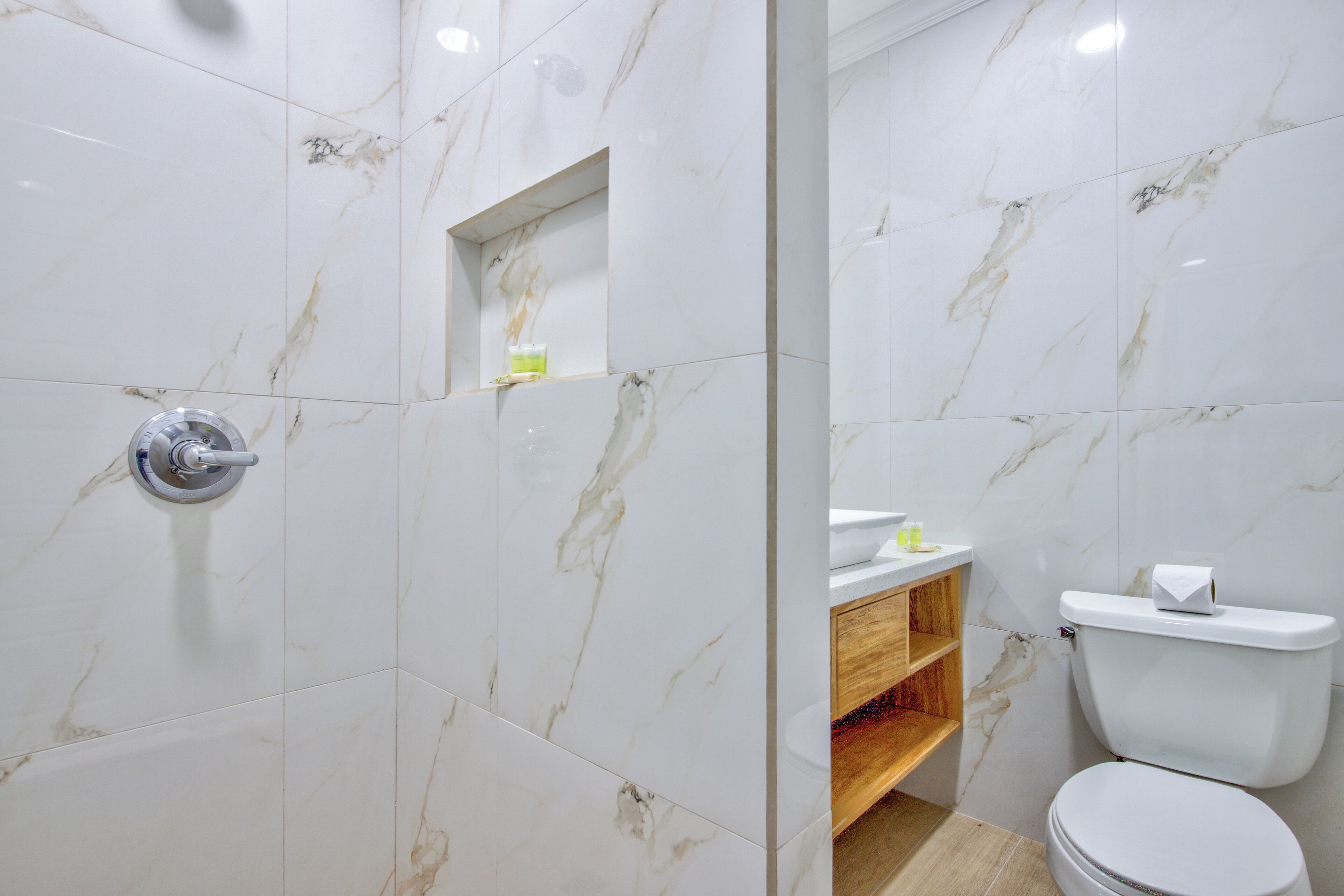 Un baño con baldosas de mármol blanco, un tocador de madera, un inodoro y una zona de ducha.