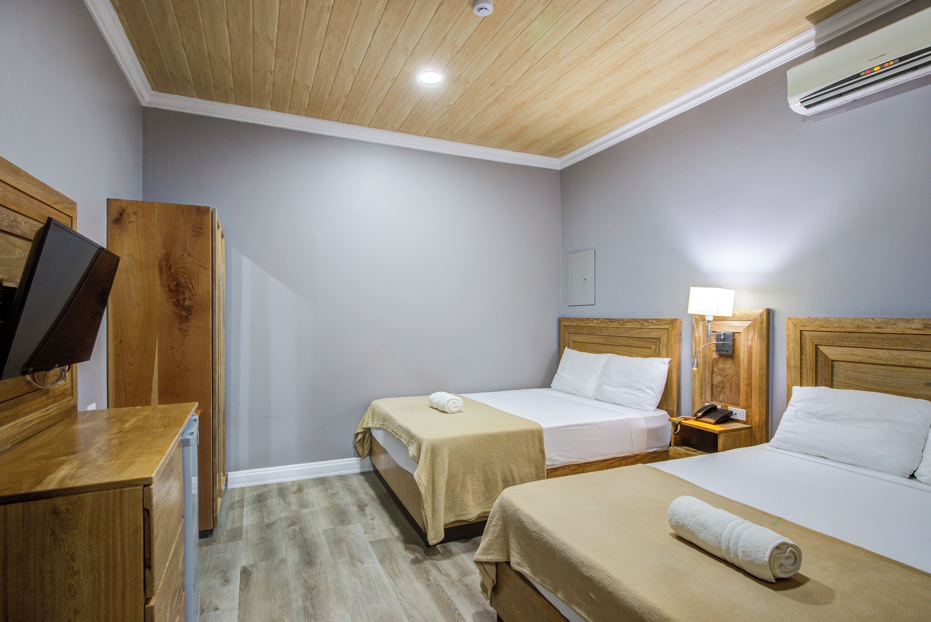 A hotel room with two beds, wooden headboards, a wooden desk, a television, and an air conditioning unit.