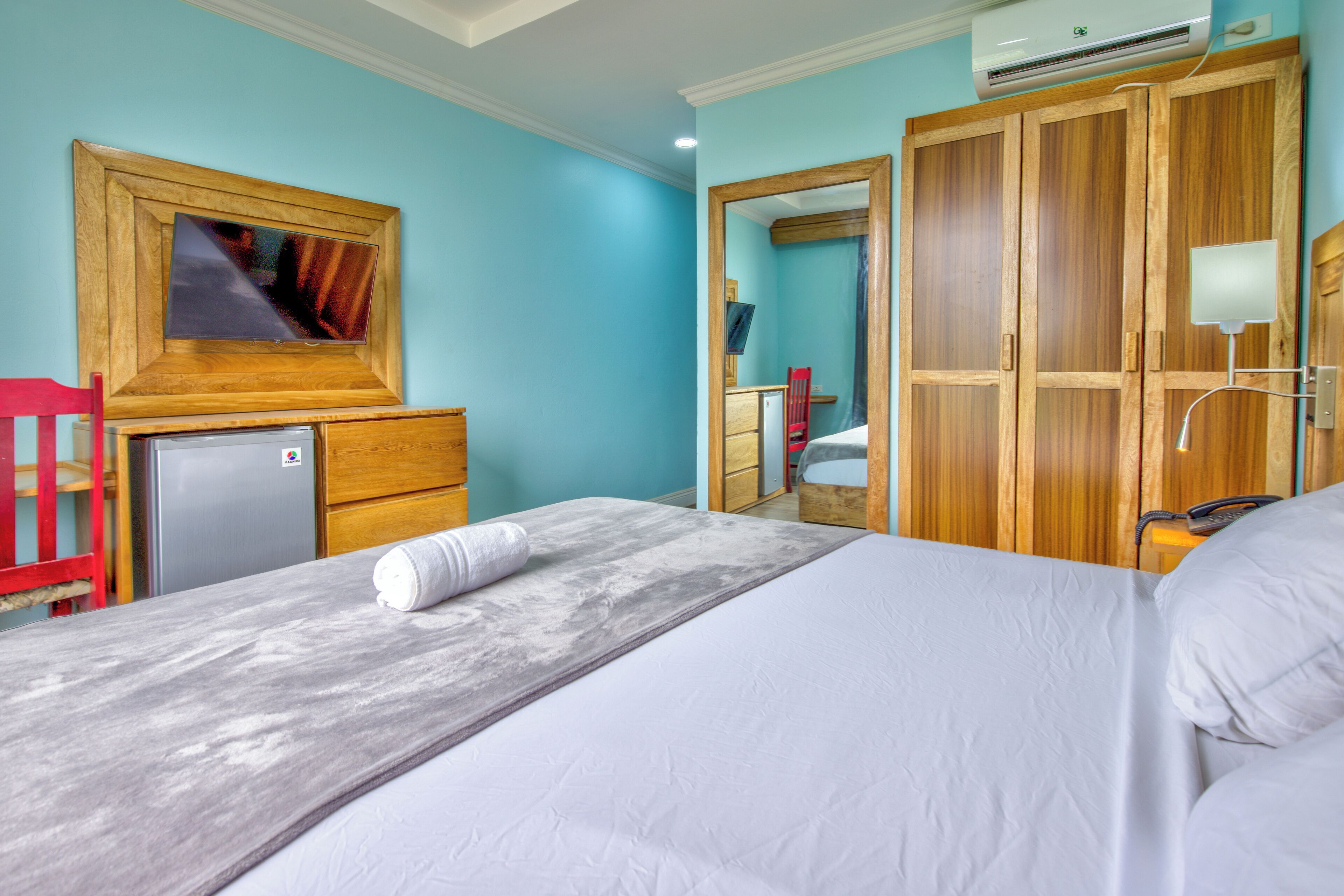 Quarto de hotel com cama, guarda-roupa de madeira, secretária pequena com cadeira, uma cadeira vermelha e um aparelho de ar condicionado.