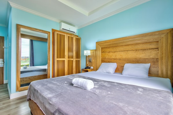 Quarto de hotel com uma cama grande, cabeceira de madeira e vista através de uma porta aberta.
