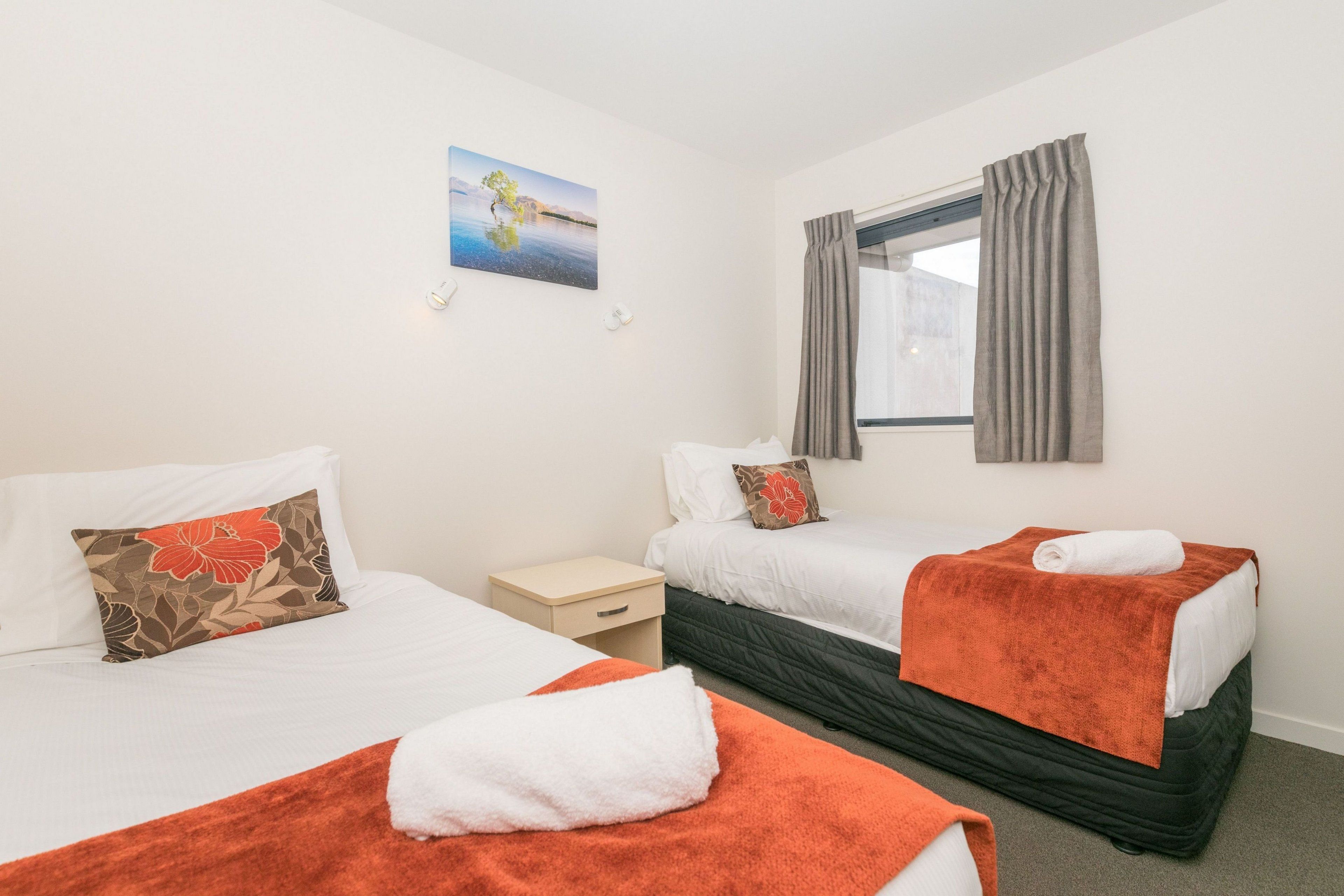 Habitación de hotel con dos camas, una mesita de noche pequeña, una ventana con cortinas y un cuadro en la pared.