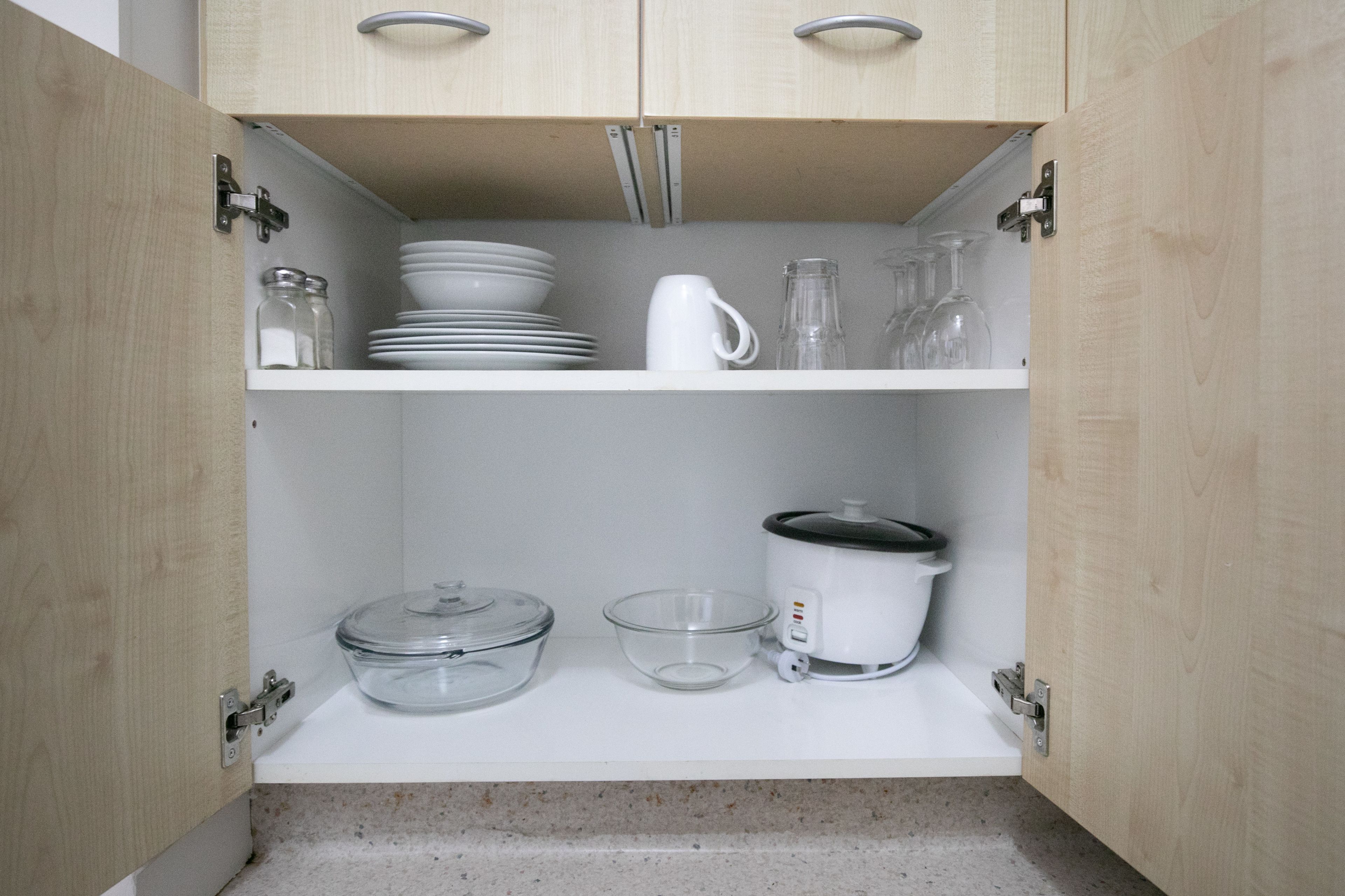 Un meuble de cuisine avec un plan de travail blanc, contenant un saladier en verre, une carafe en verre transparent, une mijoteuse blanche et une pile d’assiettes blanches.