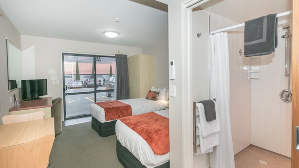 A hotel room with two beds, a desk, a TV, a shower, and a view of the parking lot. - Bella Vista Motel Wanaka (Wanaka)