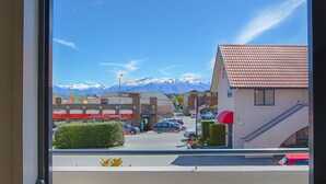 A view through a window of a building with a parking lot, a red-roofed building, and snow-capped mountains in the background. - Bella Vista Motel Wanaka (Wanaka)