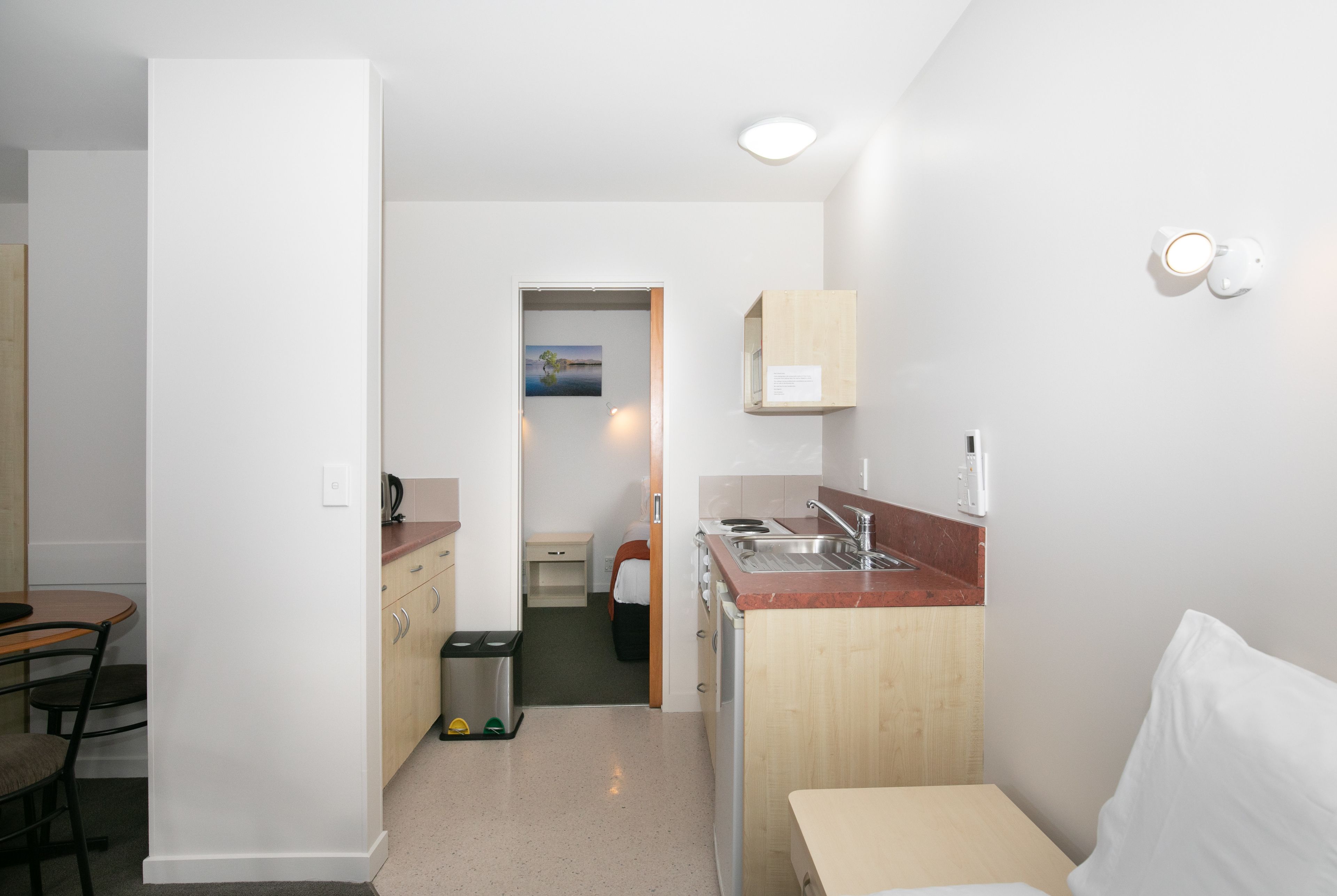 A compact kitchen with a sink, stove, and wooden cabinets. There is a small dining area with a table and chairs. A trash bin is visible near the kitchen entrance.