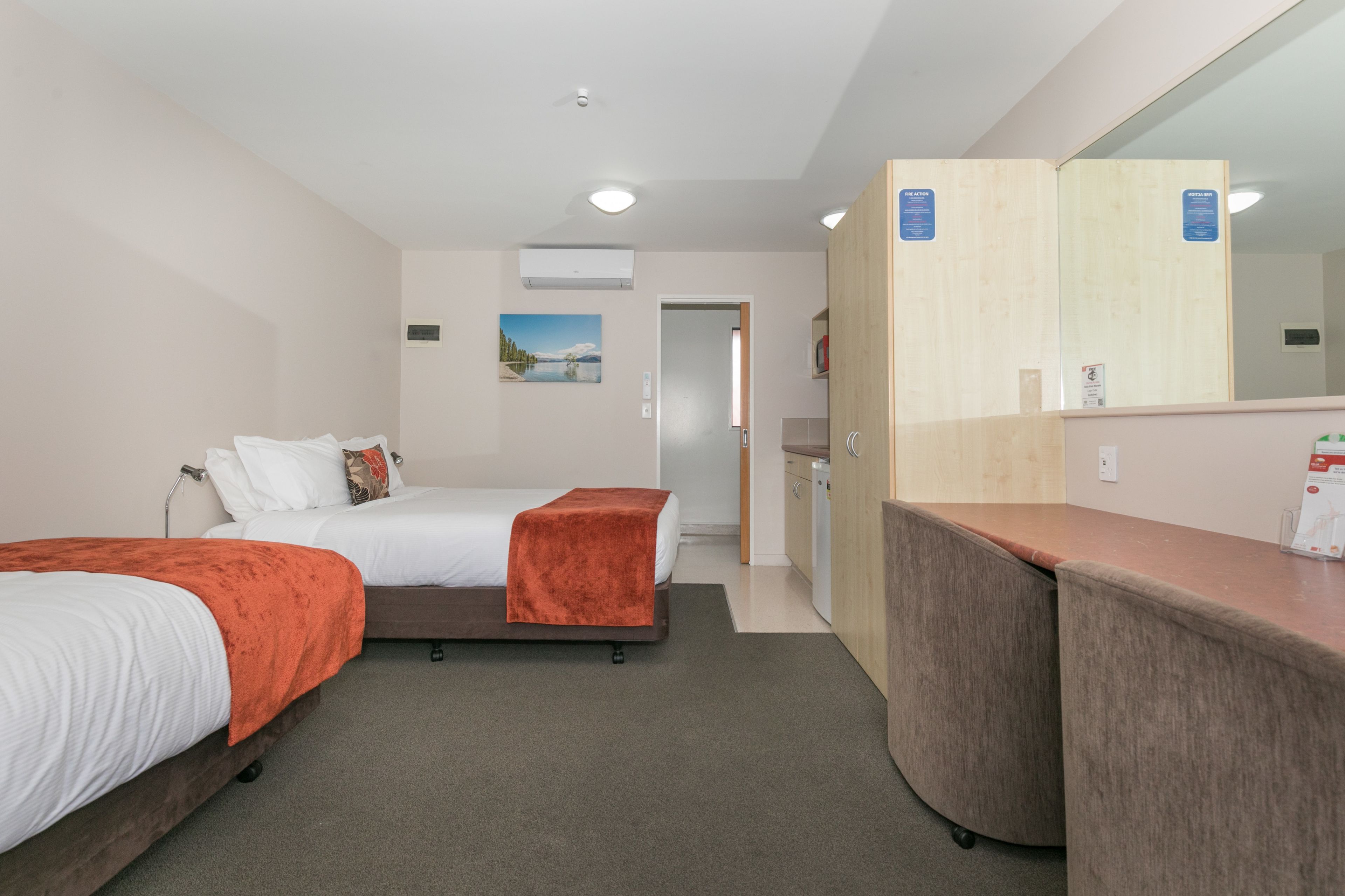 A hotel room with two beds, a desk, a mirror, and a small kitchenette.