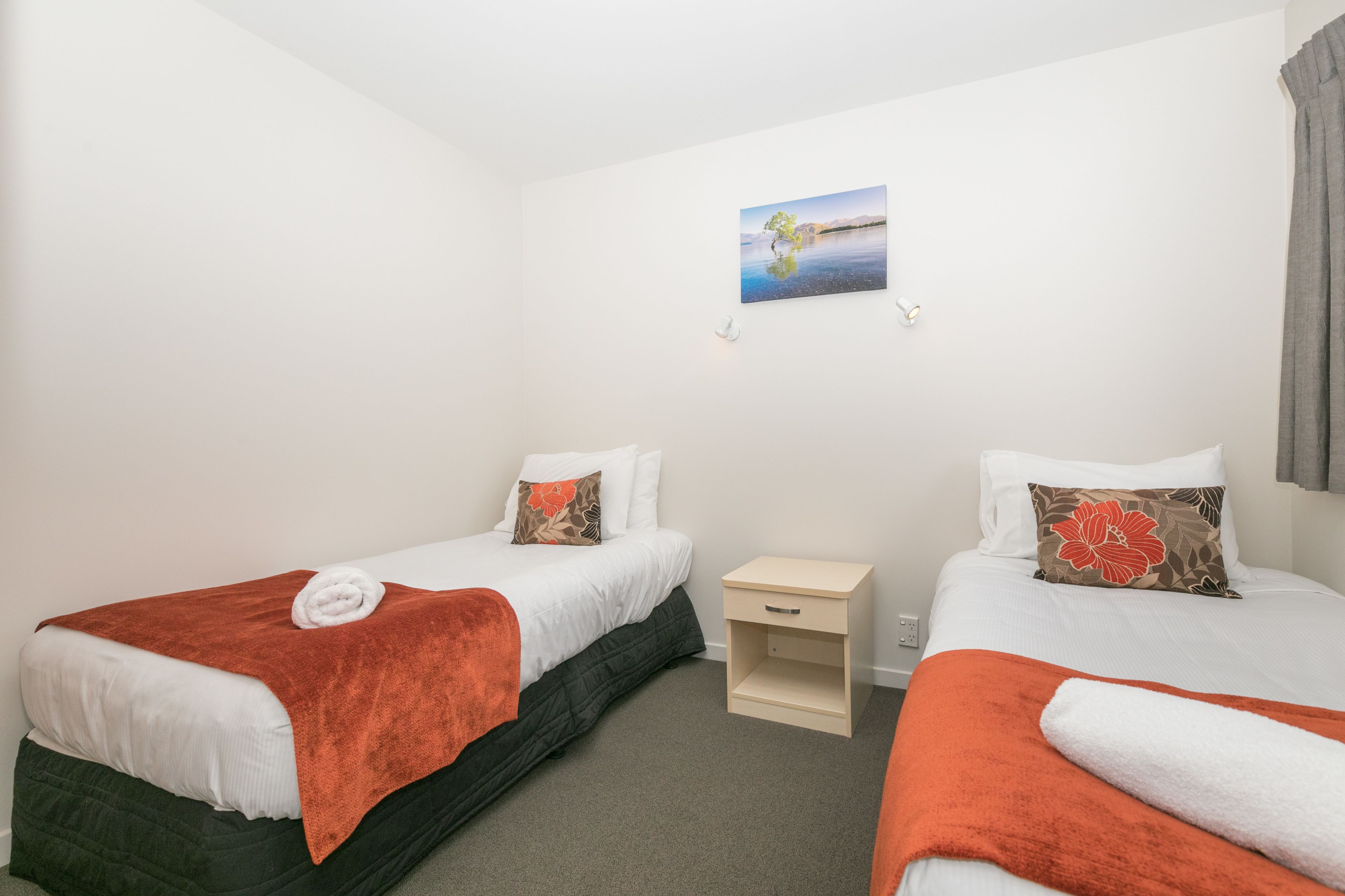 A hotel room with two single beds, each with a white duvet and orange throw blankets. There is a small bedside table with a lamp, a wall-mounted picture, and a window with curtains.