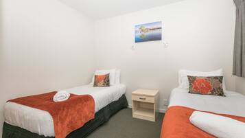 A hotel room with two single beds, each with a white duvet and orange throw blankets. There is a small bedside table with a lamp, a wall-mounted picture, and a window with curtains.