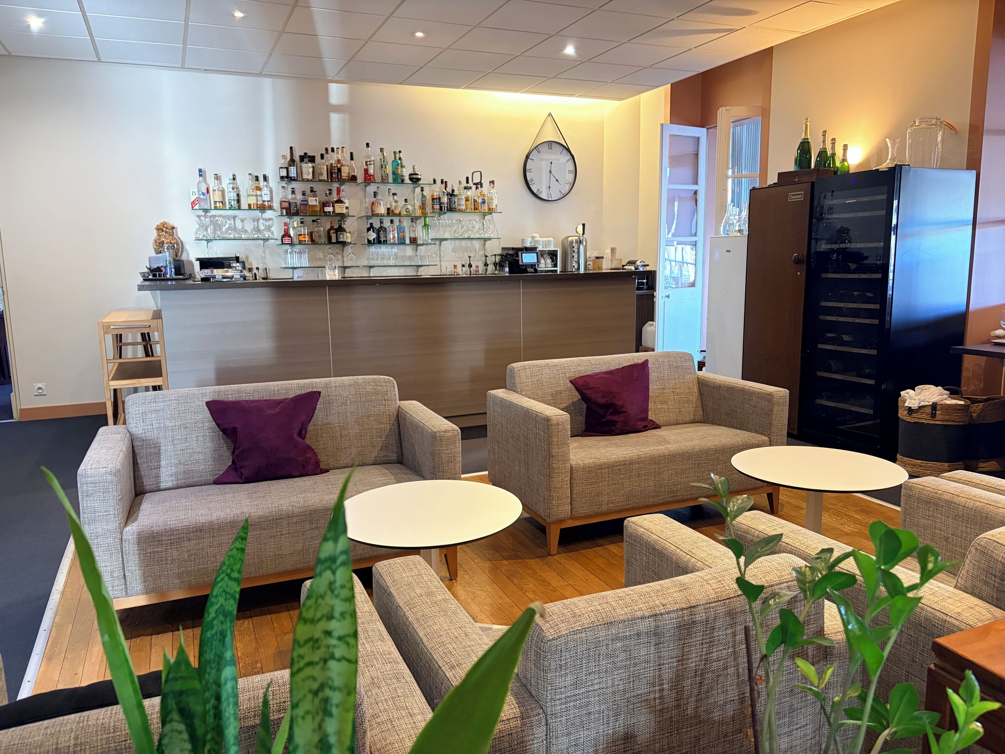 A bar area with a clock, sofas, and tables, and a refrigerator with bottles.