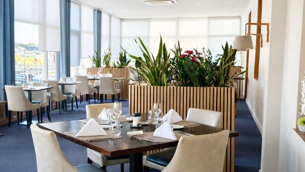 A modern dining area with tables set for two, surrounded by tall plants and a view of the city. - Grand Hotel (Mayenne)