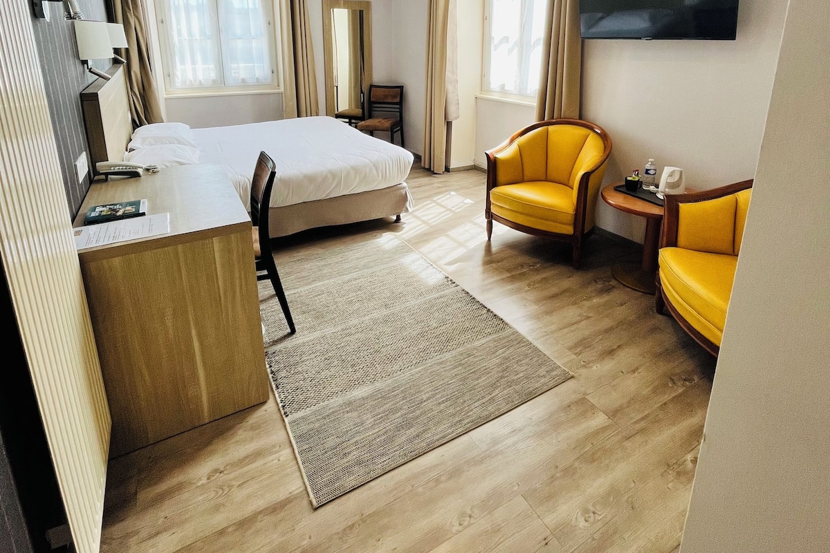 A hotel room with a bed, desk, two yellow armchairs, a TV, and a window with curtains.