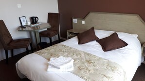 A hotel room with a bed, two pillows, a bedside table with a kettle, a chair, a small table with a phone, and a framed picture. - Logis La Rose des Vents (Volvic)