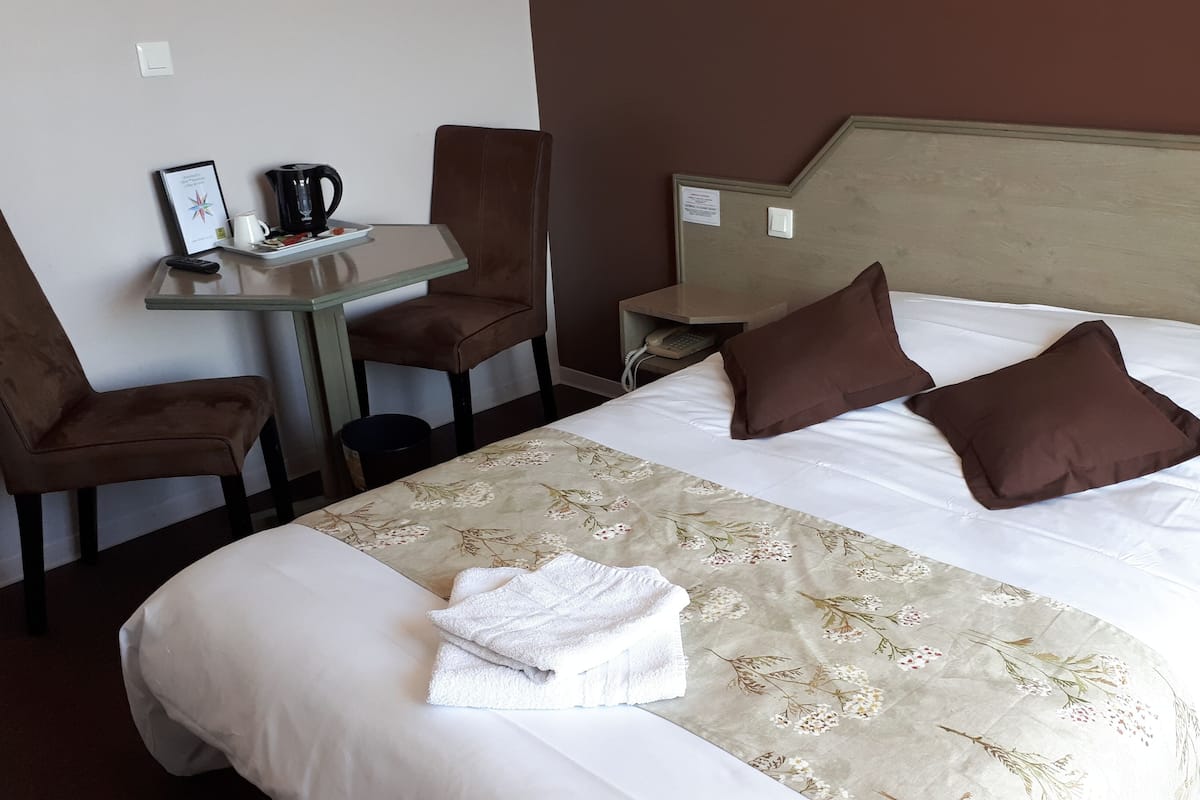 A hotel room with a bed, two pillows, a bedside table with a kettle, a chair, a small table with a phone, and a framed picture.