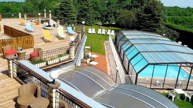 A rooftop deck with lounge chairs, a glass-enclosed pool, and a view of the surrounding greenery. - Logis La Rose des Vents (Volvic)