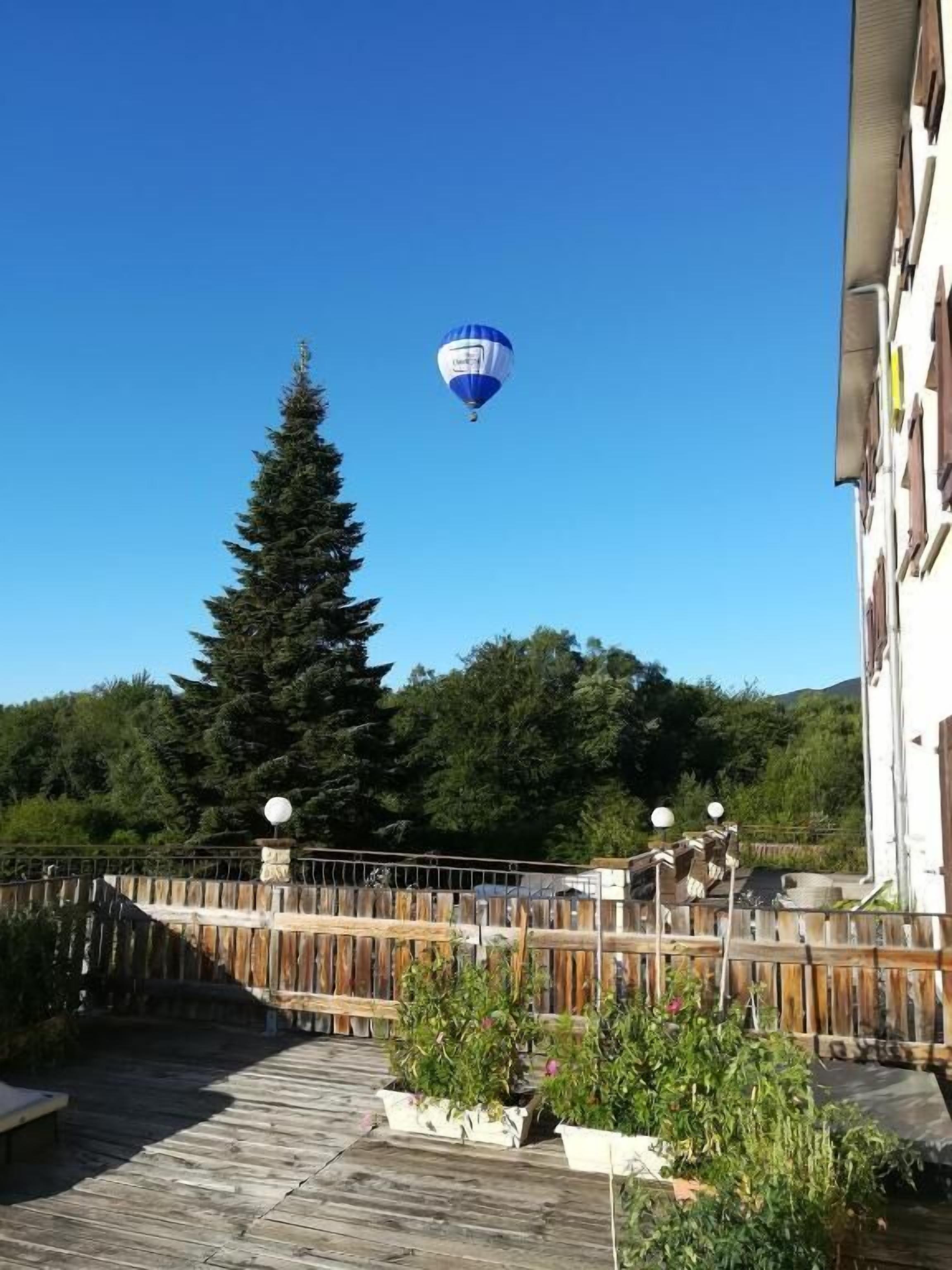 Un ballon à air chaud dans le ciel, au-dessus d’une terrasse en bois ornée de plantes en pot, avec une clôture en bois et un bâtiment en arrière-plan.