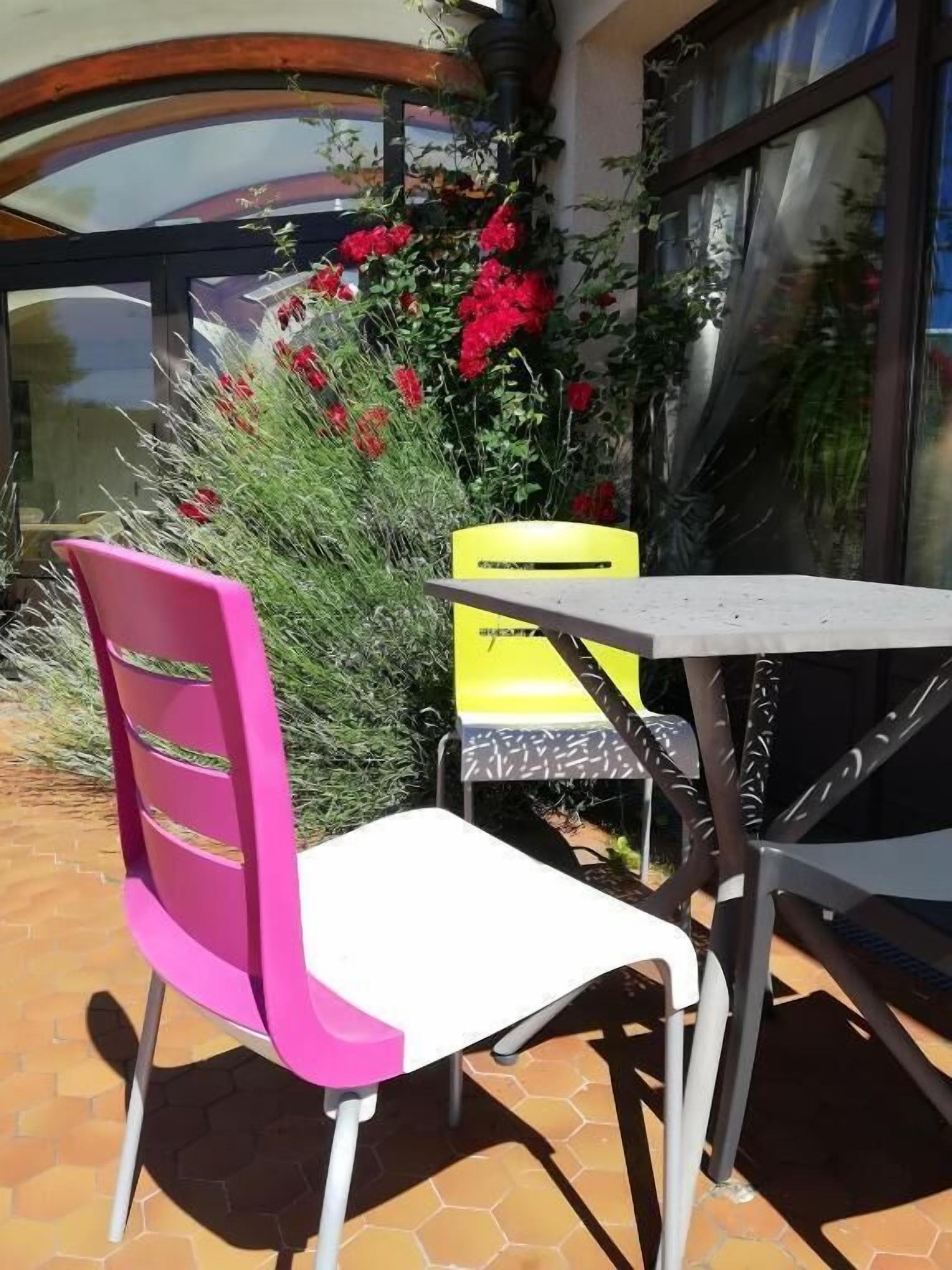 An outdoor patio with a table and a chair, surrounded by plants and a window.