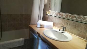 A bathroom with a beige tiled wall, a white sink, a glass shelf with toiletries, and a towel hanging on the wall.