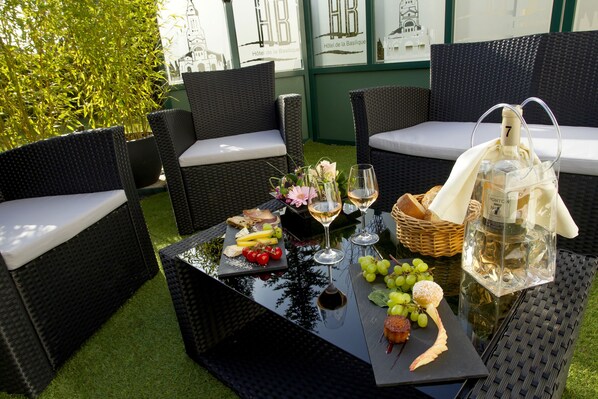 An outdoor seating area with wicker furniture, a glass table with a bottle of champagne, and a basket of bread. - Hotel & Restaurant de la Basilique (Albert)