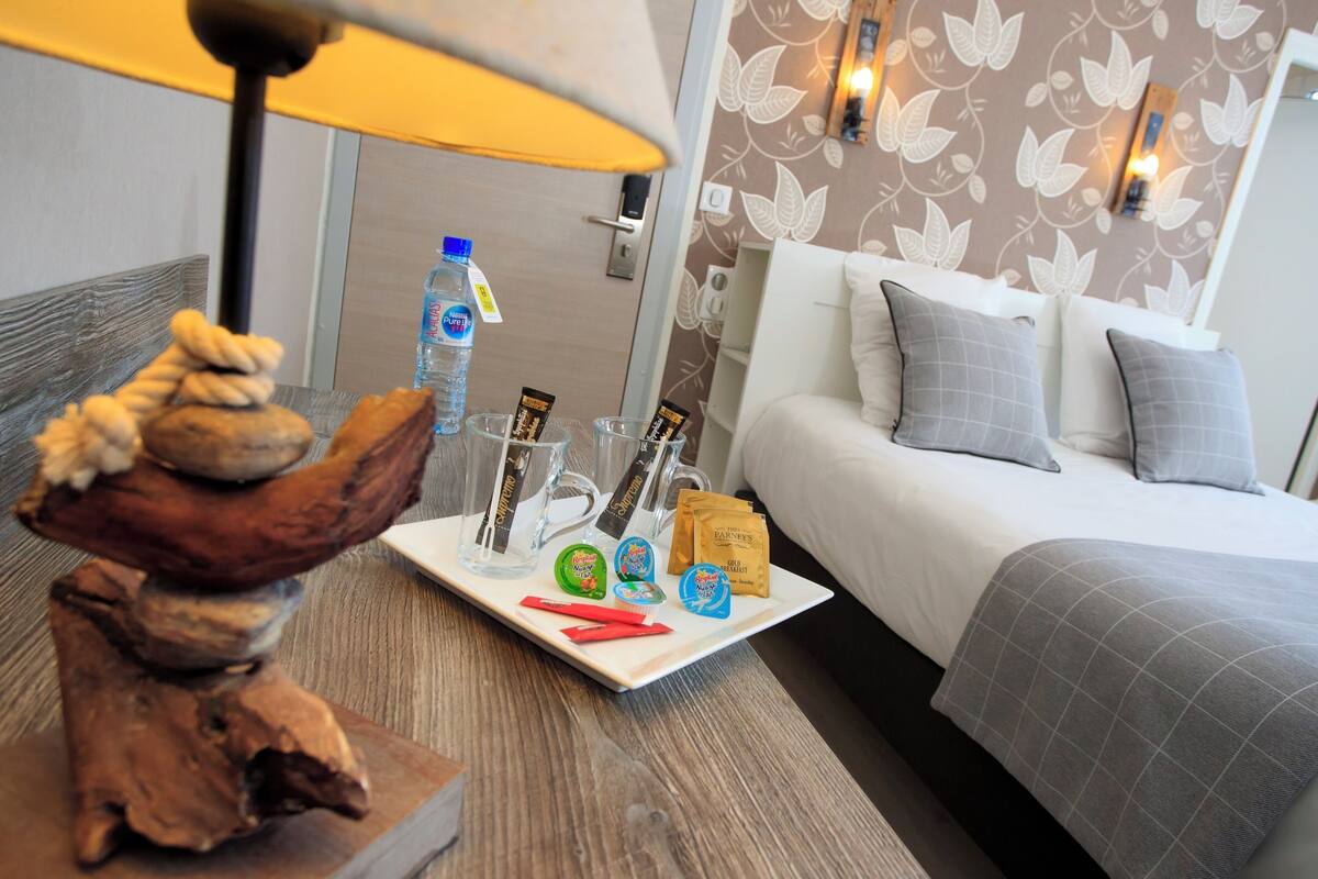 Ein Hotelzimmer mit einem Bett, einem Nachttisch mit Lampe, einer Ablage mit Snacks, einer Wasserflasche und einer dekorativen Holzskulptur.