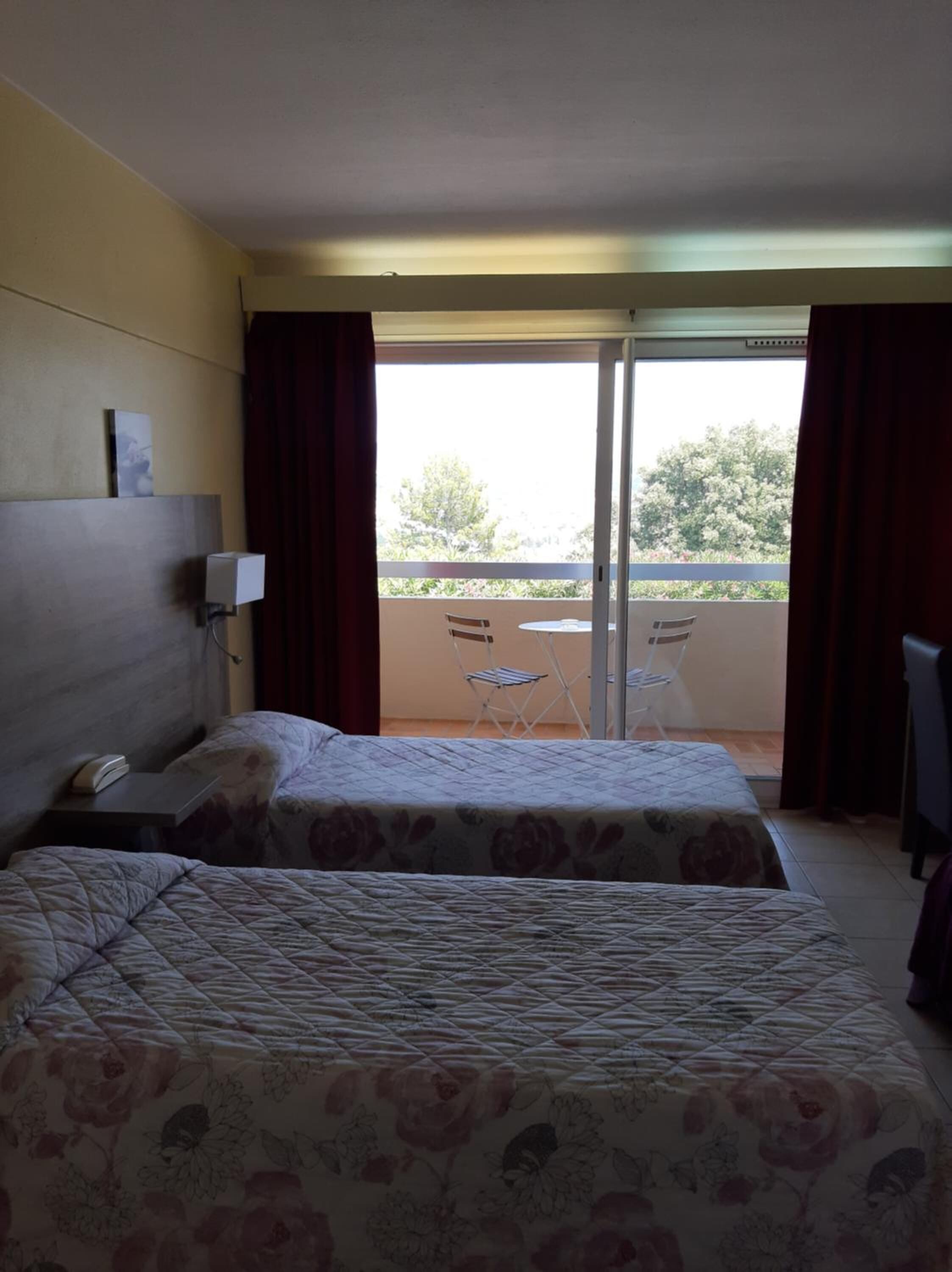 A hotel room with two beds, a small table, a chair, and a balcony with a table and chairs.