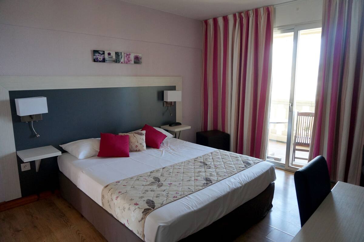 Quarto de hotel com uma cama grande, mesas de cabeceira com luminárias, uma mesinha e vista para o exterior através de uma porta deslizante.