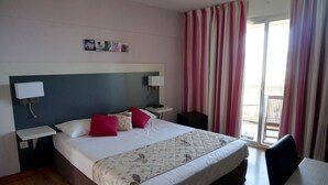 A hotel room with a large bed, bedside tables with lamps, a small table, and a view of the outside through a sliding door.