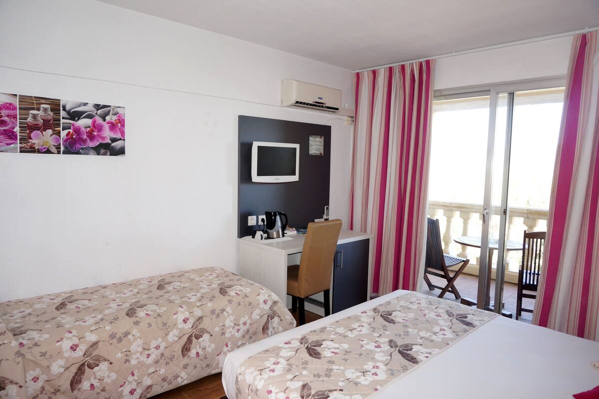 Quarto de hotel com duas camas, uma escrivaninha com cadeira, televisão e varanda com mesa e cadeiras.