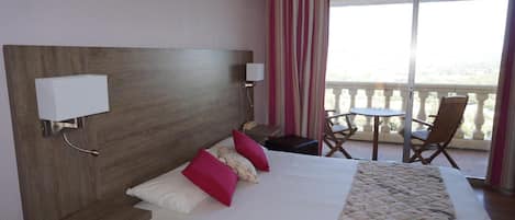 A hotel room with a large bed, two bedside tables, a nightstand, a small table with chairs, and a balcony with a view.
