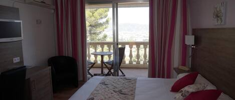 Habitación de hotel con cama, escritorio, silla, pequeña mesa, televisión y balcón con cortinas.