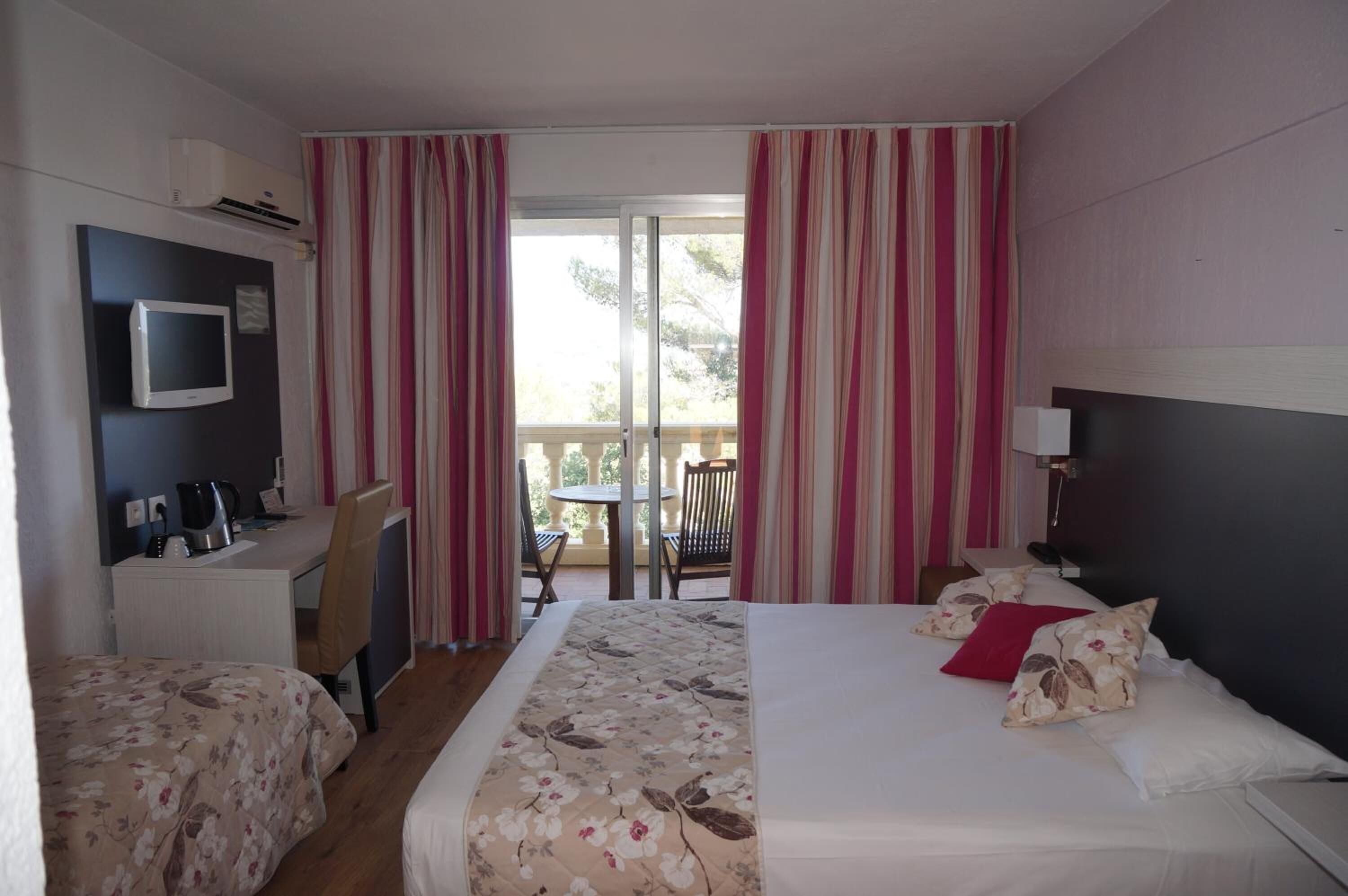 A hotel room with a bed, a desk with a chair, a television, and a balcony with striped curtains.