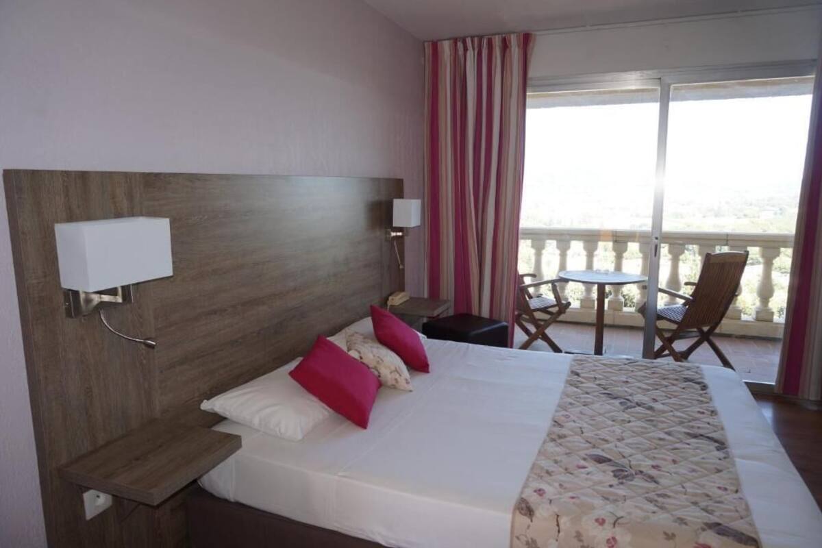 Quarto de hotel com cama, duas mesas de cabeceira, abajur, cadeira, mesa pequena e varanda com vista.