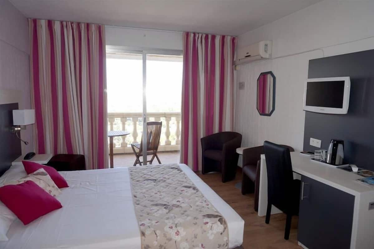 Quarto de hotel com cama, escrivaninha, cadeira, televisão e varanda com cortinas.