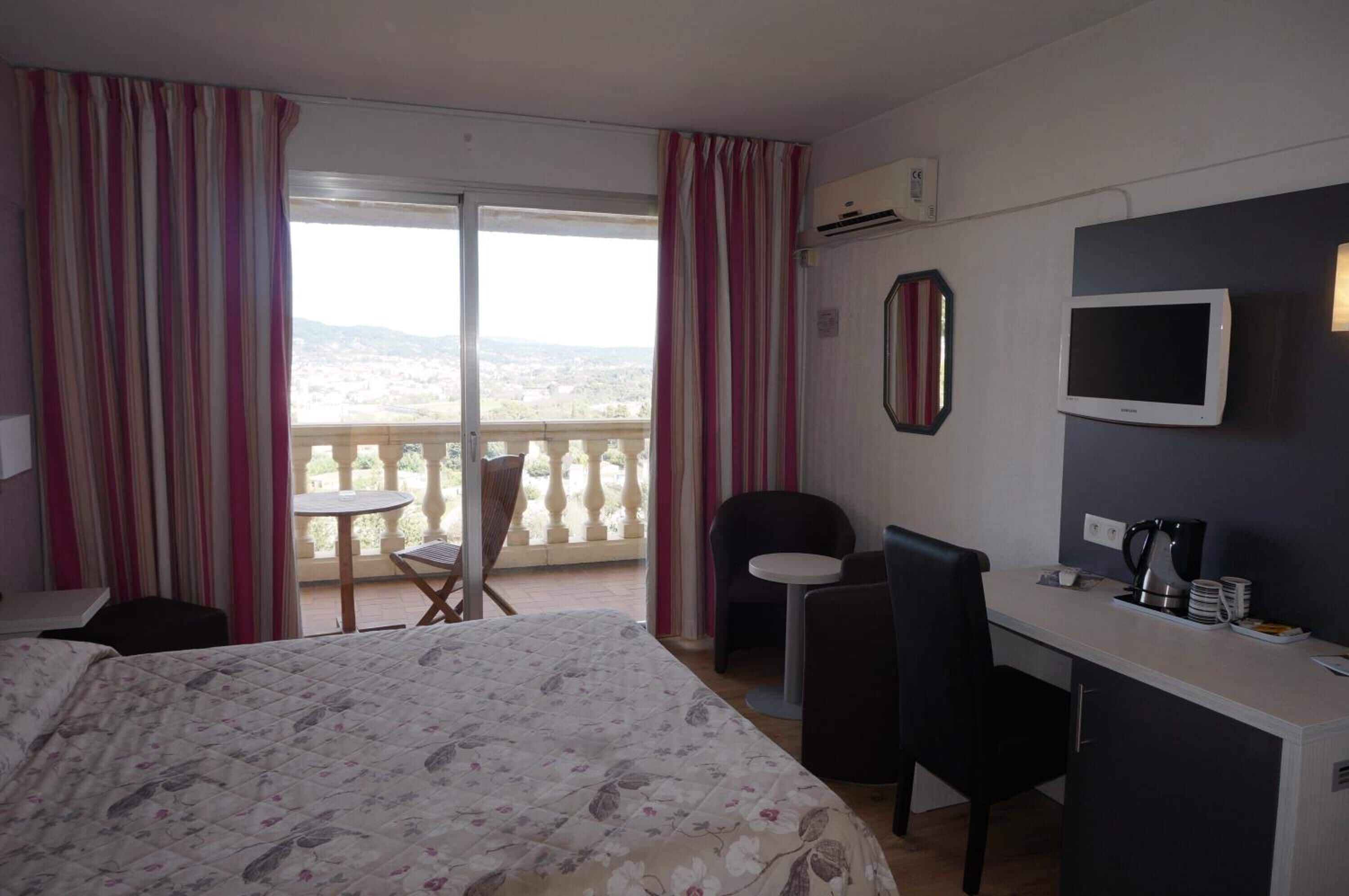 A hotel room with a bed, a desk, a chair, a small table, and a balcony with curtains.