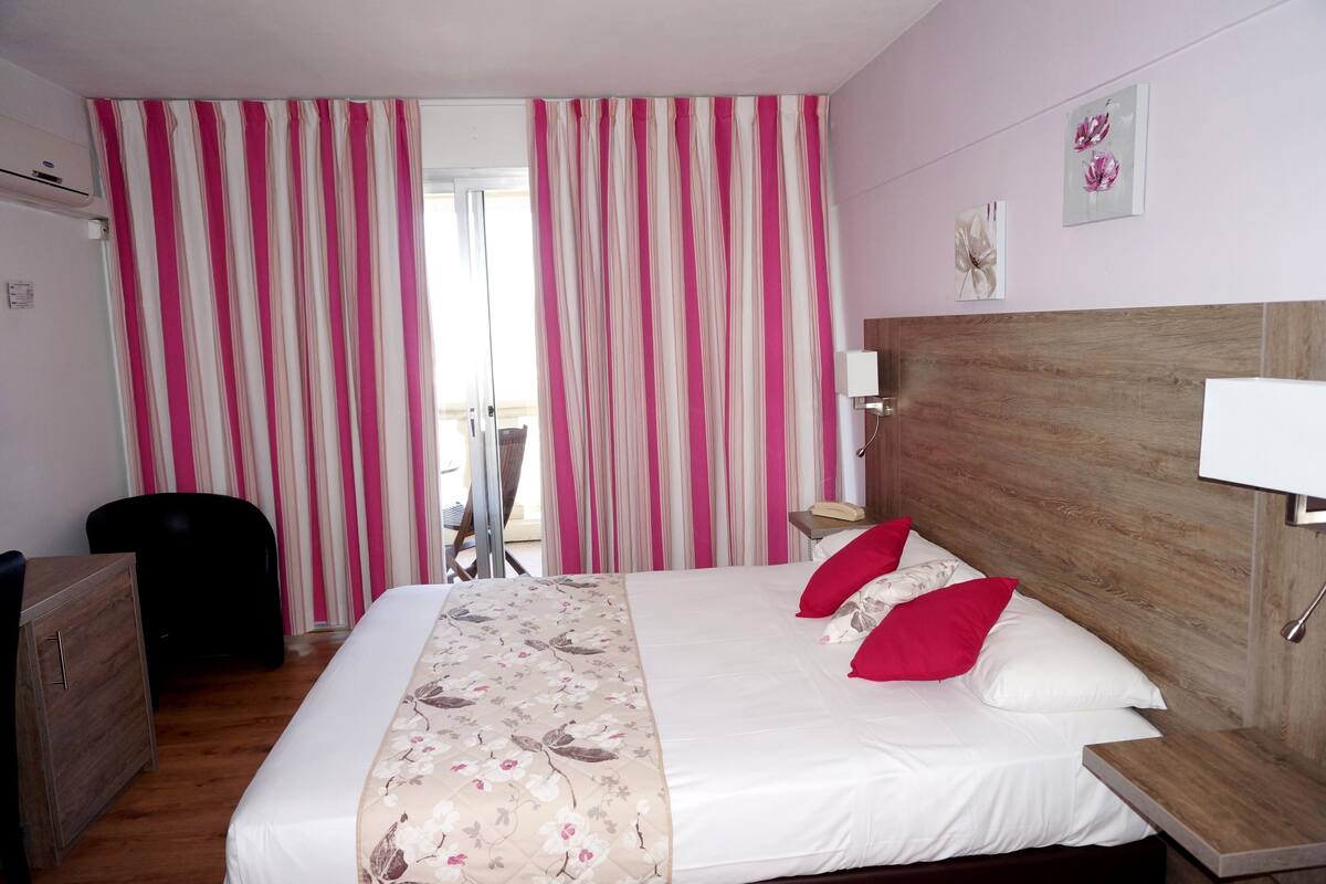 Quarto de hotel com cama, cabeceira de madeira, escrivaninha, cadeira e janela com cortinas.