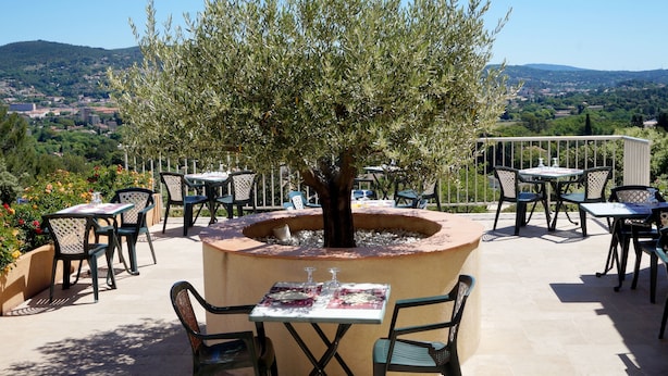 An outdoor patio with tables and chairs, a central tree, and a scenic view of a town and hills in the background.
