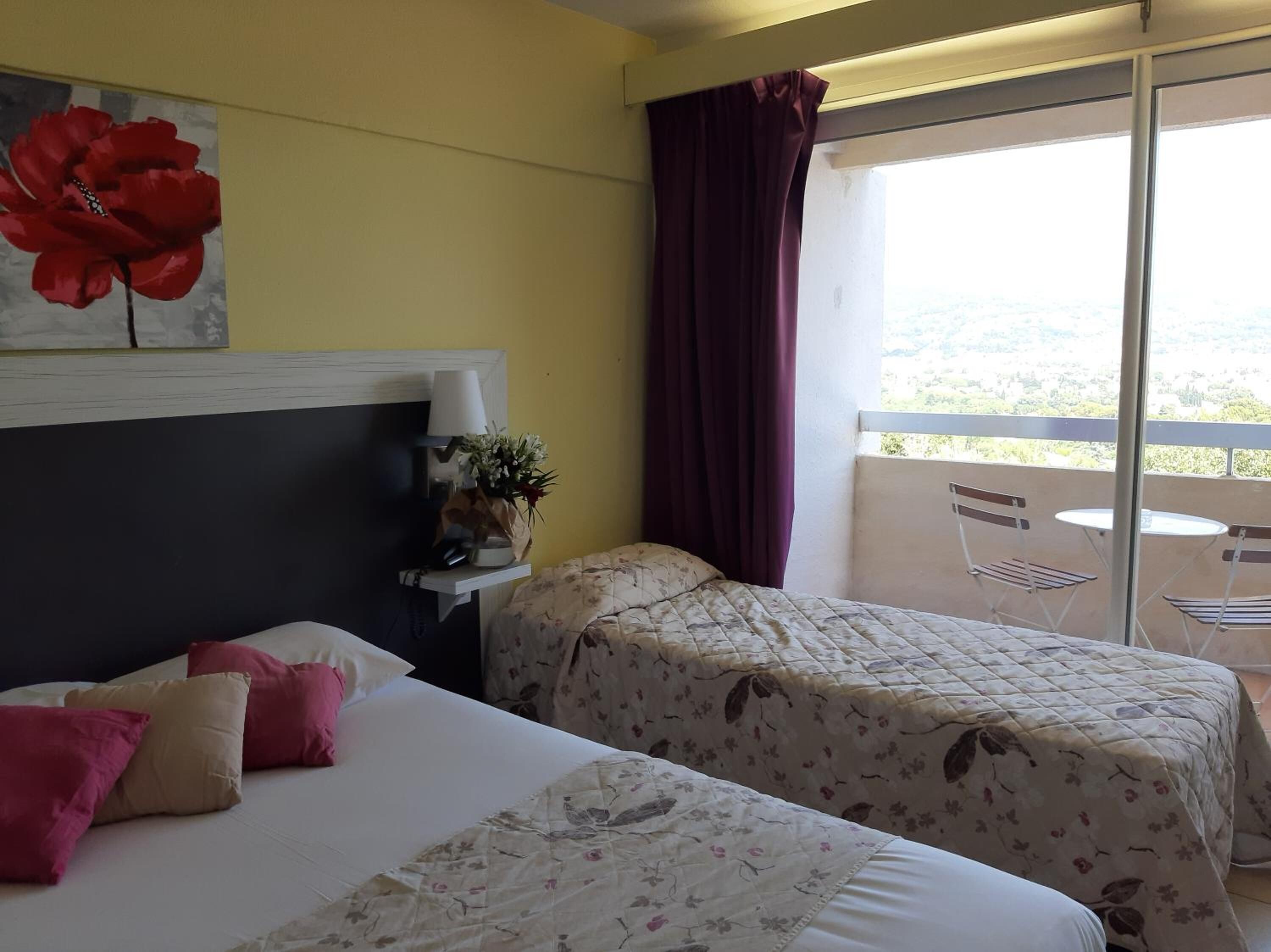 Habitació d'hotel amb dos llits, una petita taula de nit, una làmpada, un vas amb flors i un balcó amb cadires i taula.