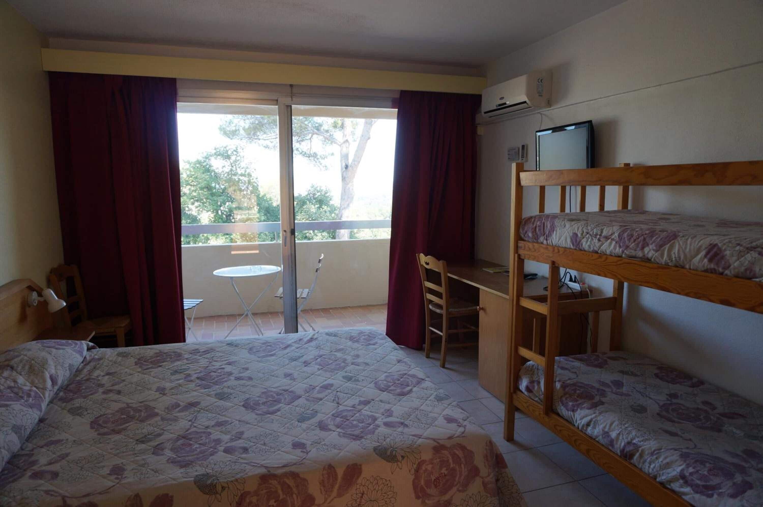 A room with a bed, a desk, and a bunk bed. There is a window with curtains, a chair, and a small table.