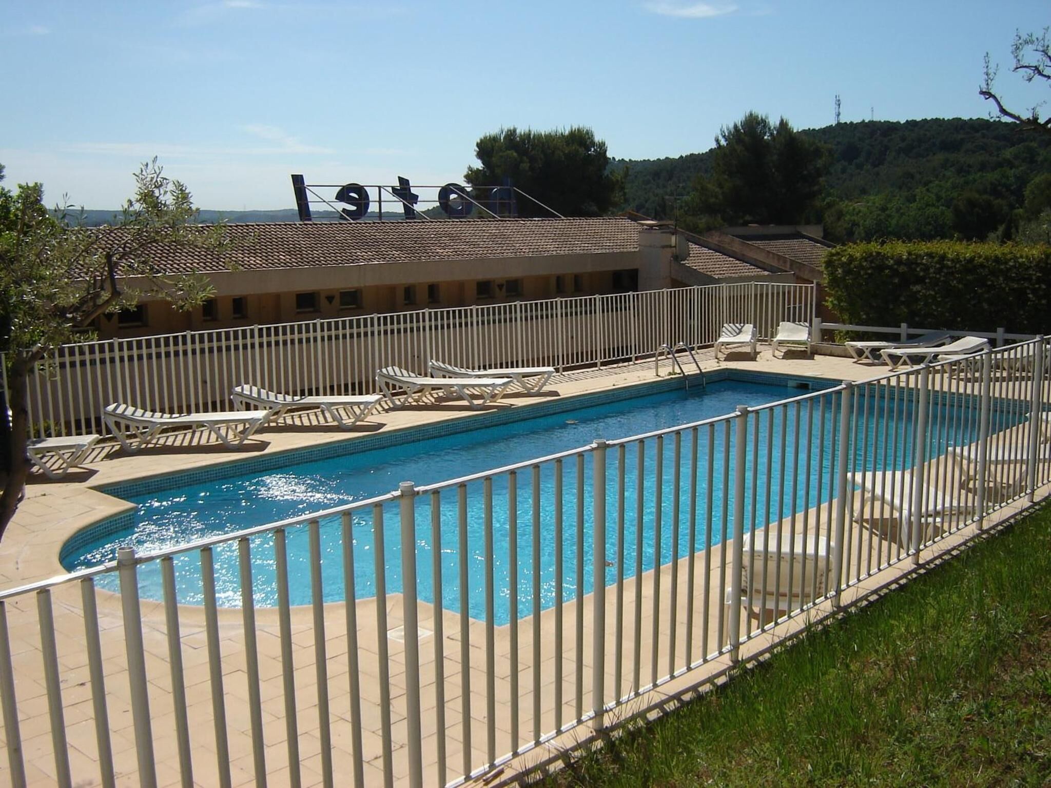 A swimming pool with a white fence, lounge chairs, and a building with a sign in the background.