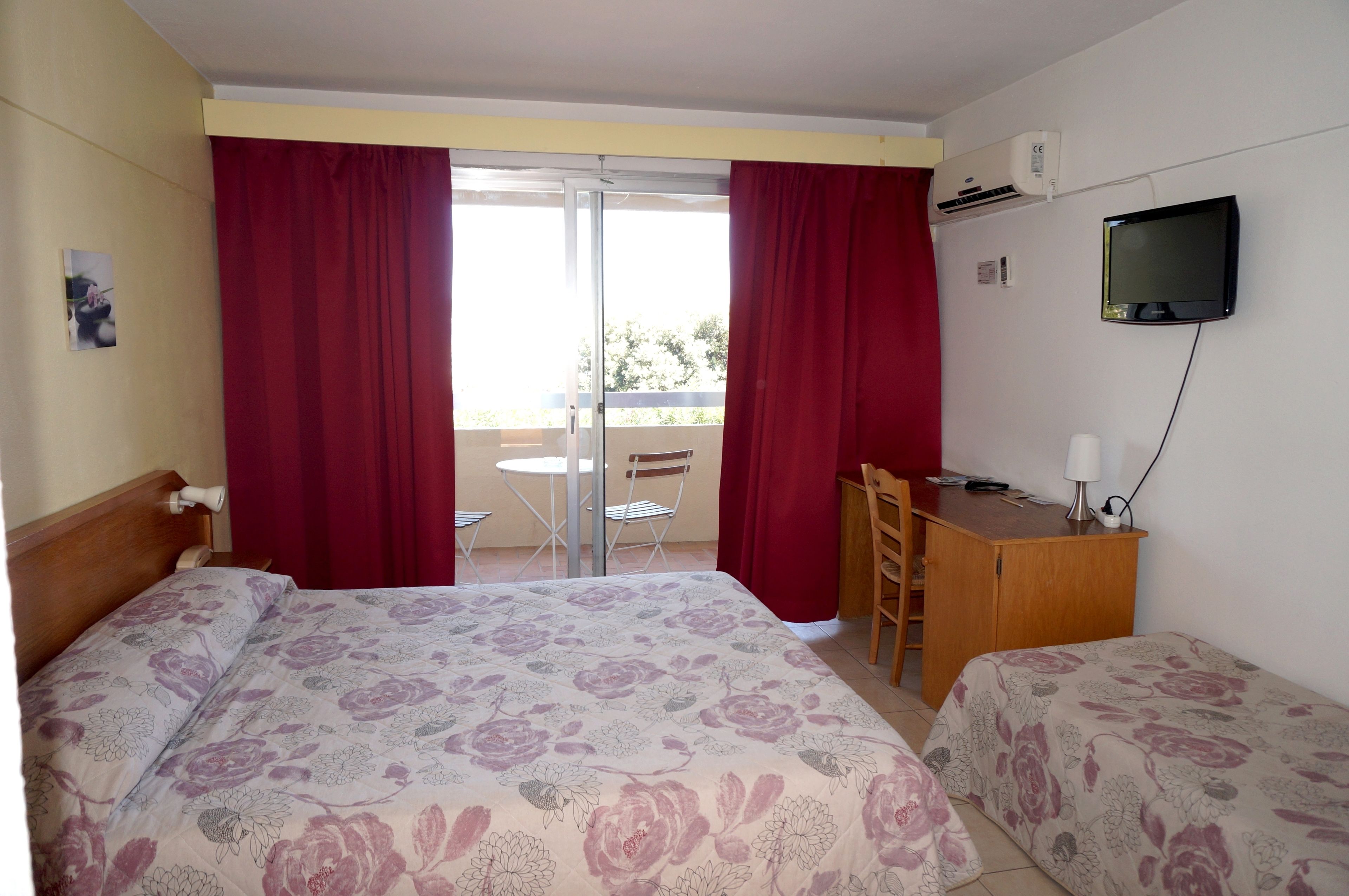 A hotel room with two beds, a desk, a chair, a television, and a balcony with red curtains.